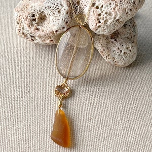 Puede incluir: Un collar de oro con un gran colgante ovalado transparente con destellos dorados. El colgante está unido a un colgante redondo más pequeño, transparente, con detalles dorados. El collar cuelga de un trozo de vidrio de mar marrón.