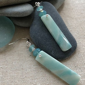 May include: A pair of dangle earrings with a light blue, rectangular, agate stone and a crystal bead. The earrings are silver toned.
