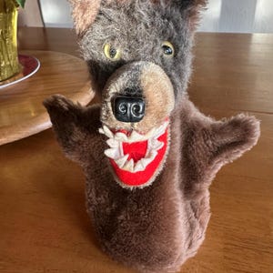 May include: A brown and grey wolf puppet with a red mouth and white teeth. The puppet has large, yellow eyes and a black nose.