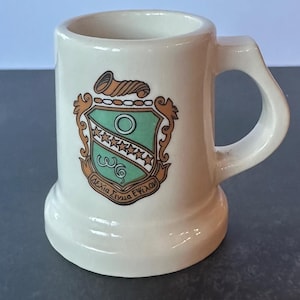 May include: A white ceramic beer stein with a green and gold crest featuring a horn, stars, and the text "Aexia Sigma Epsilon".