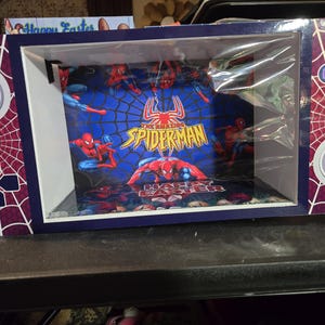 Op de afbeelding: Een decoratieve doos die is ontworpen om op een videogameconsole te lijken, met een Spider-Man-thema. De doos heeft een blauw-wit kleurenschema met een Spider-Man-afbeelding op de voor- en zijkanten en de tekst "Happy Easter".
