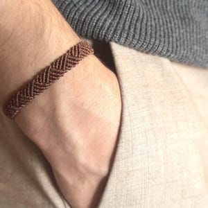 Könnte beinhalten: Ein braunes Makramee-Armband mit einem gewebten Zickzackmuster. Das Armband wird am Handgelenk getragen, mit einem grauen Pullover und einer beigen Hose im Hintergrund. Das Armband ist etwa 1,3 cm breit.