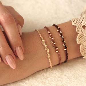 Könnte beinhalten: Vier Armbänder mit goldenen Perlen auf einem hellbraunen Hintergrund. Die Armbänder bestehen aus dünnem, geflochtenem Band in Beige, Braun und Dunkelbraun.