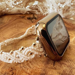 Könnte beinhalten: Ein goldfarbenes Smartwatch-Gehäuse mit einem cremefarbenen Makramee-Armband. Das Armband weist ein komplexes Flechtmuster mit kleinen, runden, metallisch goldenen Perlen auf, die in die Kordel eingearbeitet sind und mit einem Schiebeknoten enden.
