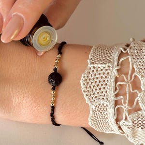 Könnte beinhalten: Ein Handgelenk mit einem schwarzen Armband mit einer goldenen Perle und einer schwarzen Lavasteinperle. Die Person trägt ein klares Fläschchen mit ätherischem Öl auf ihr Handgelenk auf.