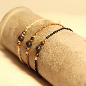 Könnte beinhalten: Drei Armbänder mit Perlen in Creme, Braun und Schwarz, jeweils mit Lavastein- und Tigeraugenperlen. Die Armbänder werden auf einer zylindrischen, strukturierten Oberfläche präsentiert, die die natürlichen Farbtöne des Schmucks hervorhebt.