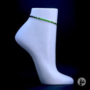 May include: A black and green beaded ankle bracelet on a white mannequin leg.