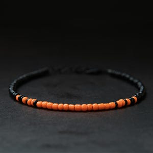 May include: A circular bracelet constructed from small, uniform beads. The design features a repeating pattern of vibrant orange beads interspersed with segments of solid black beads, creating a high-contrast, simple, and linear aesthetic.