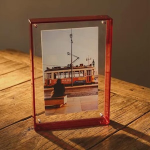 Puede incluir: Un marco de fotos rectangular de acrílico rojo que muestra una fotografía de un tranvía antiguo. El marco tiene una parte delantera y trasera de acrílico transparente, sujetas por pequeños remaches plateados. La imagen muestra a una persona sentada en un banco.