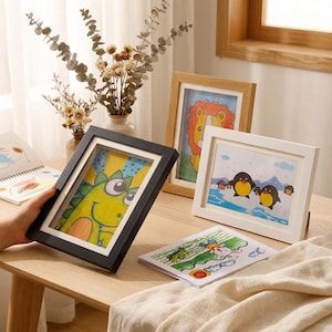 May include: Three framed children's drawings on a wooden table. The frames are black, white, and light brown. The drawings feature a green dinosaur, a lion, and penguins. A drawing of a dragon and a drawing of a rabbit are also visible.