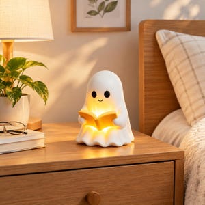 May include: A white ghost-shaped night light with a smooth finish, featuring two black circular eyes and a small curved mouth. The ghost is holding an open yellow book that emits a warm, soft glow from within its structure.