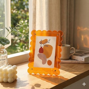 Könnte beinhalten: Ein orangefarbener Acryl-Bilderrahmen mit gewelltem Rand. Der Rahmen enthält einen Druck mit abstrakten Formen in Orange, Braun und Rosa. Der Rahmen steht auf einer Holzoberfläche.