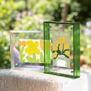 Op de afbeelding: Twee heldere acryl fotolijsten, één met een groene rand, tonen bloemenkunstprints. Het linker frame bevat een geel en groen bloemendesign, terwijl het rechter frame een "Tulip Garden" print met gele tulpen heeft.