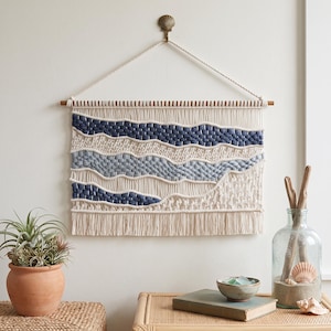 May include: A macrame wall hanging featuring cream-colored cotton cord with woven horizontal bands in shades of dark navy and light blue. The piece is suspended from a wooden dowel by a braided cord and finished with a fringe at the bottom edge.