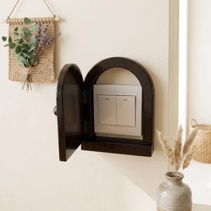 May include: A dark brown wooden arched wall cover featuring a hinged door that opens to reveal a dual light switch. The frame is constructed with a smooth, polished finish and is mounted directly over a standard rectangular electrical switch plate.