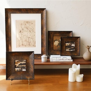 May include: An assortment of framed art pieces in various sizes, featuring abstract and floral prints. The frames have a textured, dark brown finish. A small ceramic jar, books, candles, and a vase are also on display.