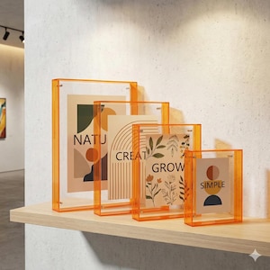 May include: Four clear acrylic picture frames with orange borders, each holding a different art print. The prints feature the words "NATU", "CREATE", "GROW", and "SIMPLE" with abstract designs in neutral colors. The frames are displayed on a light-colored wooden shelf.