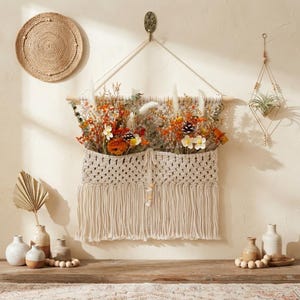 May include: A cream-colored macrame wall hanging with a pocket containing dried flowers in shades of orange, yellow, and white, accented with pinecones and greenery. The piece features a wooden dowel, hanging cord, and long fringe at the bottom.