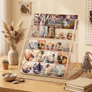 Könnte beinhalten: Ein klarer Acryl-Displayständer mit mehreren Regalen, gefüllt mit bunten Sammelkarten und Ansteckern. Der Ständer steht auf einem Holztisch, im Hintergrund eine Vase mit Trockenblumen und eine kleine Sukkulentenpflanze.