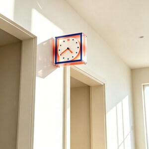 Op de afbeelding: Een vierkante wandklok met een witte ronde wijzerplaat, blauwe stip-uurmarkeringen en oranje wijzers. De klok is gevat in een transparant frame met blauwe randen en is bevestigd aan een witte muur tussen twee deuropeningen in een lichte kamer.