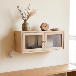 Puede incluir: Un armario de pared de madera clara con dos puertas que tienen paneles de vidrio estriado y pequeños pomos redondos de madera. Un cable de alimentación blanco con enchufe cuelga del lado izquierdo. El armario está montado en una pared lisa.