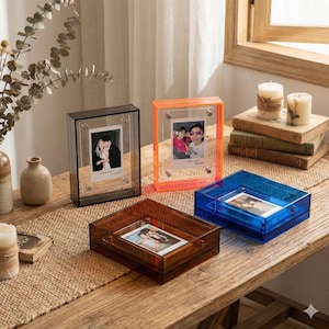 Puede incluir: Una colección de marcos de fotos y cajas de exhibición de acrílico de colores. Los marcos son en tonos negro, naranja y azul, cada uno con una fotografía. Las cajas son en tonos marrón y azul. Los artículos están colocados sobre una mesa de madera.