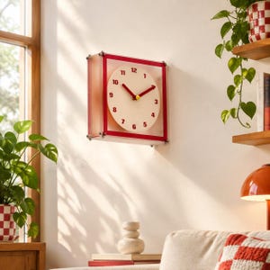 May include: A square wall clock featuring a white circular face with red numerals from 1 to 12. The clock has red hour and minute hands with a small yellow centre cap. It is encased in a transparent frame with red borders and silver hardware.