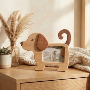 May include: A wooden dog-shaped photo frame, featuring a picture of a small white dog. The frame is light brown with darker brown ears and tail. It's displayed on a wooden surface, with a neutral background.