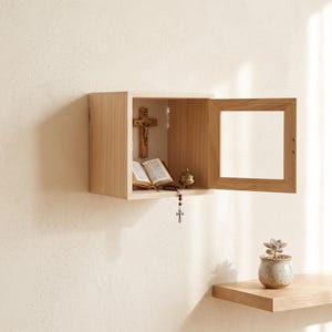 Op de afbeelding: Een lichtgekleurde houten wandkast met een glazen deur. Binnenin de kast is een houten crucifix aan de achterwand bevestigd, vergezeld van een open boek en een kleine metalen houder, met een rozenkrans die over de rand naar beneden hangt.