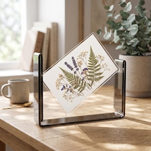 May include: A rectangular clear acrylic floating frame held by a dark gray U-shaped stand. Inside the frame, pressed dried flowers and green fern leaves are arranged on a white background, featuring purple lavender sprigs and small white blossoms.