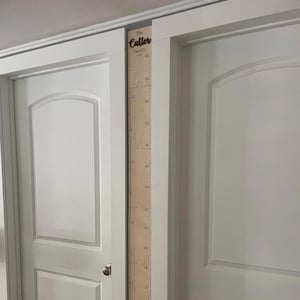 Może przedstawiać: Drewniany wykres wzrostu z oznaczeniami pomiarowymi i tekstem "The Cutter Family". Wykres jest zamontowany na ramie drzwi pomiędzy dwiema białymi drzwiami. Wykres wzrostu ma jasnobrązowy kolor.