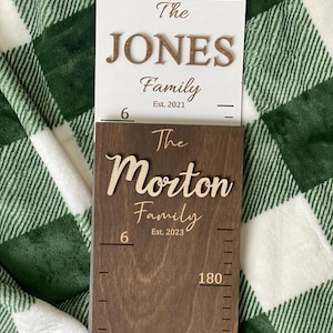 May include: A wooden growth chart with a brown stain. The chart has a white sign at the top that reads "The JONES Family Est. 2021". The chart also has the text "The Morton Family Est. 2023" and a ruler that goes up to 180 cm.