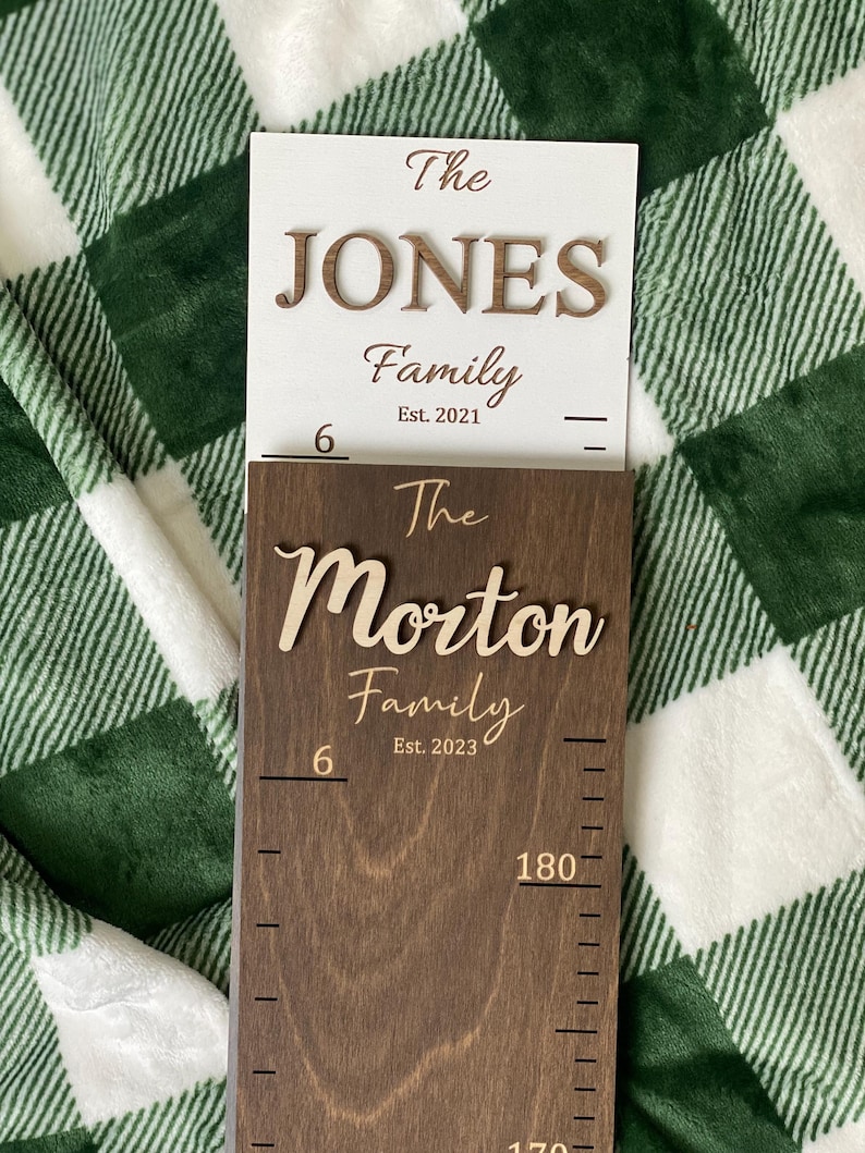 May include: A wooden growth chart with a brown stain. The chart has a white sign at the top that reads "The JONES Family Est. 2021". The chart also has the text "The Morton Family Est. 2023" and a ruler that goes up to 180 cm.