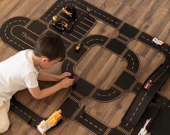May include: A black and tan wooden toy road playmat with a child playing with toy cars. The playmat has a road design with dashed lines, crosswalks, and the name "Thomas" printed on it. Several toy vehicles are on the road.