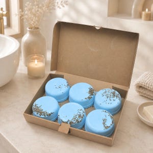 Puede incluir: Un conjunto de seis bombas de baño redondas de color azul claro dispuestas en una caja de cartón marrón. Cada bomba de baño cilíndrica y lisa está cubierta con hierbas botánicas secas, creando un contraste texturizado sobre la superficie azul.