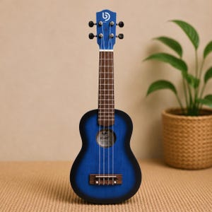 Könnte beinhalten: Eine blaue Sopran-Ukulele mit dunkelblauem Sunburst-Finish, braunem Griffbrett und vier weißen Saiten. Die Kopfplatte zeigt ein weißes Logo und das Schallloch enthält ein sichtbares Etikett. Das Instrument hat einen Steg und vier Stimmwirbel.