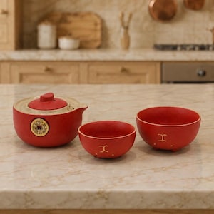 May include: A red ceramic teapot with a gold-coloured coin emblem on the side and two matching red bowls featuring a small gold-coloured symbol. The teapot has a rounded lid and a spout, while the bowls are circular with small feet at the base.