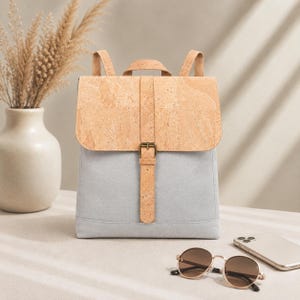 May include: A backpack featuring a light grey fabric body and a tan cork material flap. The bag includes a vertical cork strap with a metal buckle closure, a top handle, and two shoulder straps extending from the back of the main compartment.