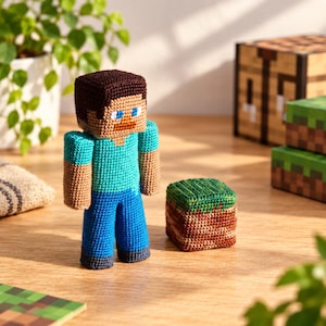 Puede incluir: Una figura de ganchillo de un personaje cuadrado con pelo castaño, camisa azul claro y pantalones azul oscuro, junto a un pequeño bloque de hierba de ganchillo cuadrado con la parte superior verde y los lados marrones, en hilo.