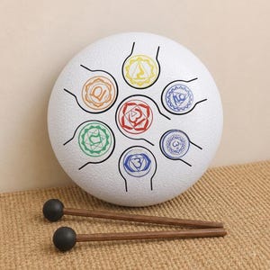 Pode incluir: Um tambor de língua de aço branco com sete recortes circulares distintos, cada um marcado com um símbolo de chakra colorido em vermelho, laranja, amarelo, verde e azul. Dois martelos de madeira com cabeças esféricas pretas estão à frente.