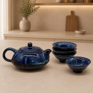 Op de afbeelding: Een keramische theeset bestaande uit een ronde theepot met deksel en vier bijpassende kleine kommen. De onderdelen hebben een diepblauw glazuur met lichtere blauwe en zwarte gespikkelde accenten, met een gladde en glanzende afwerking aan de buitenkant.