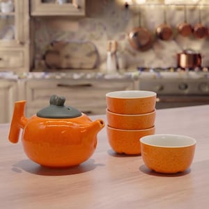 Könnte beinhalten: Eine leuchtend orangefarbene Keramik-Teekanne mit dunkelgrauem Deckel und Griff, begleitet von einem Stapel aus drei passenden orangefarbenen Schalen und einer einzelnen orangefarbenen Schale, alle mit strukturierter Außenseite und glatter weißer Innenseite.