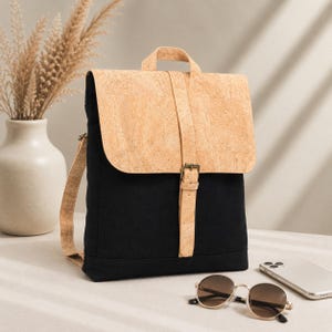 May include: A backpack featuring a black fabric body and a light tan cork flap with a matching cork top handle and shoulder straps. The flap is secured by a vertical cork strap with a metal buckle closure, providing a contrast between the dark textile and the natural cork texture.