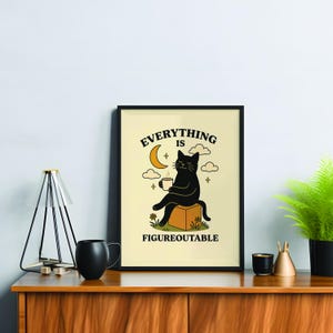 Peut inclure: Impression d'art encadrée avec un chat noir assis sur une boîte, tenant une tasse de café, avec le texte "Everything is Figureoutable". L'illustration a un fond crème avec une lune, des nuages et des étoiles.