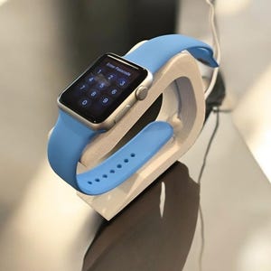 Op de afbeelding: Een zilveren Apple Watch met een lichtblauw sportbandje wordt getoond op een witte standaard. Het horloge-scherm toont een numeriek toetsenbord met de tekst "Enter Passcode".
