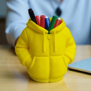 Peut inclure: Un porte-stylos jaune vif en forme de sweat à capuche, avec une poche avant et une capuche. Le porte-stylos est rempli de stylos et de marqueurs colorés, dont noir, rouge, bleu et vert. Le porte-stylos est posé sur une surface en bois.