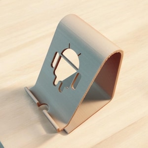 May include: A light brown wooden phone stand with an Android logo cut out. The stand is angled and has a smooth surface.