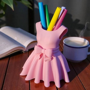 Peut inclure: Un organiseur de bureau rose en forme de robe contient des stylos colorés. Le support a un nœud à la taille et une jupe évasée. Une tasse de café et un livre ouvert sont en arrière-plan.