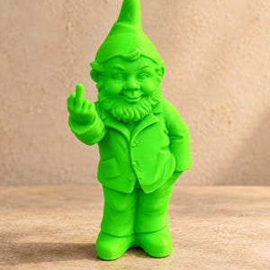 Puede incluir: Una figura de gnomo de jardín de color verde neón brillante que lleva un sombrero puntiagudo y una chaqueta. El gnomo tiene una barba poblada y se muestra con el brazo derecho levantado, extendiendo el dedo corazón en un gesto.