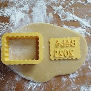 Peut inclure: Deux emportes-pièces jaunes sur une surface farinée. Un emporte-pièce est en forme de rectangle avec des bords festonnés. L'autre emporte-pièce est en forme de rectangle avec les mots "2023 Bebe" en relief.